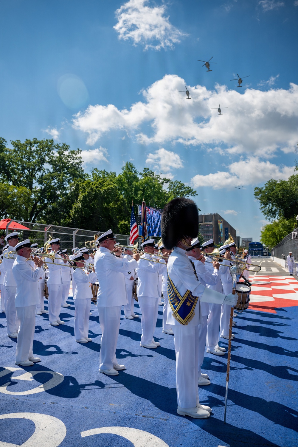 A military marching band in white dress uniforms performs outdoors on a blue and red decorated surface, with an American flag and color guard visible in the background. A drum major wearing a tall black bearskin hat stands prominently in the foreground holding a ceremonial mace. Multiple helicopters are visible flying in formation overhead in a partly cloudy blue sky.