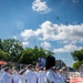 A military marching band in white dress uniforms performs outdoors on a blue and red decorated surface, with an American flag and color guard visible in the background. A drum major wearing a tall black bearskin hat stands prominently in the foreground holding a ceremonial mace. Multiple helicopters are visible flying in formation overhead in a partly cloudy blue sky.