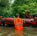 Indiana National Guard personnel are conducting flood rescue operations in a flooded waterway surrounded by dense green trees. A soldier wearing an orange high-visibility vest labeled 'Indiana National Guard' is wading in floodwater alongside rescue swimmers in red drysuits and helmets, maneuvering a large inflatable rescue boat equipped with a Mercury outboard motor. The scene depicts an active emergency flood response operation.