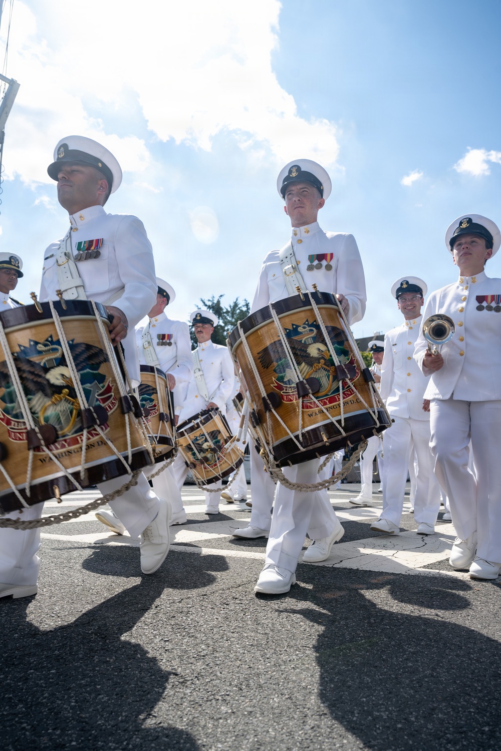 A U.S. Navy marching band is performing in an outdoor parade, with drummers in the foreground carrying ornately decorated bass drums bearing the insignia of 'The United States Navy Band, Washington D.C.' The musicians are dressed in formal white Navy dress uniforms with white caps, medals, and white shoes. The low-angle photograph captures the procession in bright daylight with a partly cloudy sky in the background.