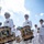 A U.S. Navy marching band is performing in an outdoor parade, with drummers in the foreground carrying ornately decorated bass drums bearing the insignia of 'The United States Navy Band, Washington D.C.' The musicians are dressed in formal white Navy dress uniforms with white caps, medals, and white shoes. The low-angle photograph captures the procession in bright daylight with a partly cloudy sky in the background.