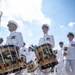 A U.S. Navy marching band is performing in an outdoor parade, with drummers in the foreground carrying ornately decorated bass drums bearing the insignia of 'The United States Navy Band, Washington D.C.' The musicians are dressed in formal white Navy dress uniforms with white caps, medals, and white shoes. The low-angle photograph captures the procession in bright daylight with a partly cloudy sky in the background.
