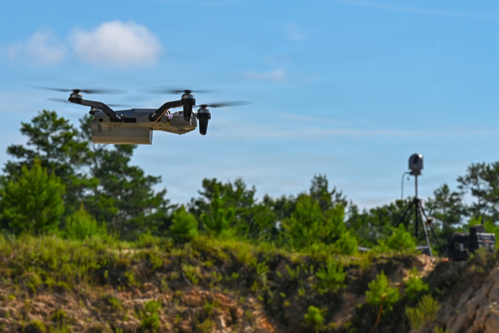 A quadcopter drone is captured mid-flight against a blue sky with scattered clouds. In the background, a tripod-mounted sensor or camera device is positioned on an elevated ridge surrounded by green vegetation. The scene appears to be an outdoor field test or demonstration environment.