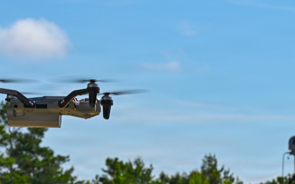 A quadcopter drone is captured mid-flight against a blue sky with scattered clouds. In the background, a tripod-mounted sensor or camera device is positioned on an elevated ridge surrounded by green vegetation. The scene appears to be an outdoor field test or demonstration environment.