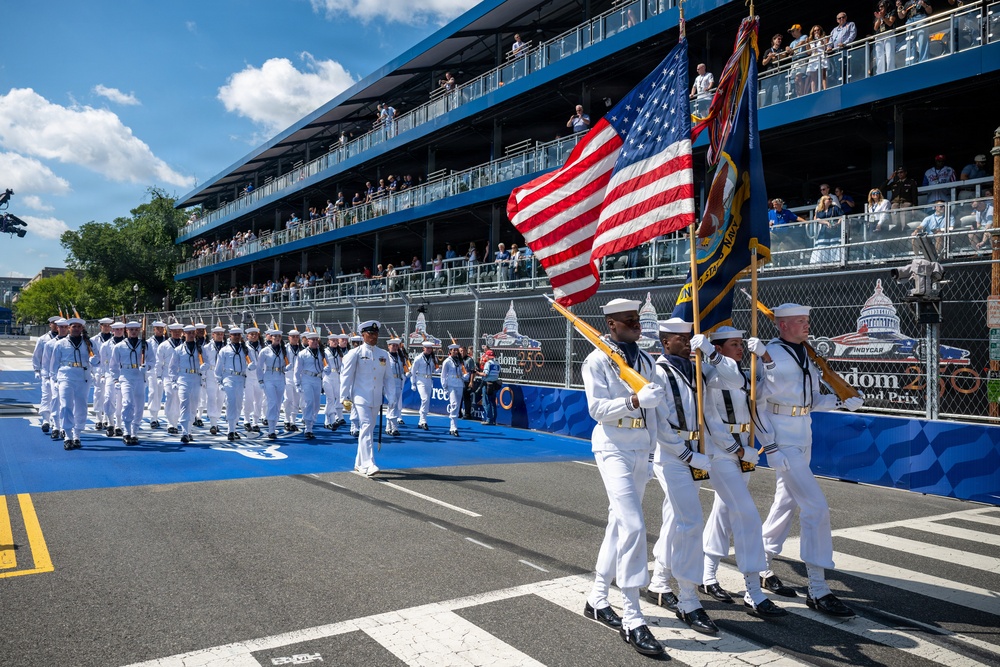 A U.S. Navy color guard and marching formation are participating in a ceremonial parade on a racing circuit, identified by signage as the 'Freedom 250 Grand Prix' IndyCar event. Sailors in dress white uniforms carry the American flag and what appears to be a Navy unit flag while spectators watch from multi-level grandstands. The U.S. Capitol building is visible in the background, confirming the location as Washington, D.C.