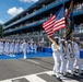 A U.S. Navy color guard and marching formation are participating in a ceremonial parade on a racing circuit, identified by signage as the 'Freedom 250 Grand Prix' IndyCar event. Sailors in dress white uniforms carry the American flag and what appears to be a Navy unit flag while spectators watch from multi-level grandstands. The U.S. Capitol building is visible in the background, confirming the location as Washington, D.C.