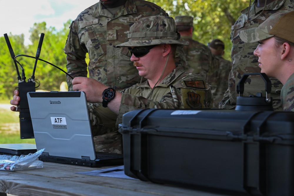 Military personnel in OCP (Operational Camouflage Pattern) uniforms are gathered around a field table operating a Panasonic Toughbook laptop labeled 'ATF Toughbook.' The central figure, wearing a boonie hat and sunglasses, appears to be connecting cables or configuring communications equipment. The setting appears to be an outdoor field exercise or training environment.