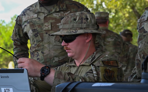 Military personnel in OCP (Operational Camouflage Pattern) uniforms are gathered around a field table operating a Panasonic Toughbook laptop labeled 'ATF Toughbook.' The central figure, wearing a boonie hat and sunglasses, appears to be connecting cables or configuring communications equipment. The setting appears to be an outdoor field exercise or training environment.