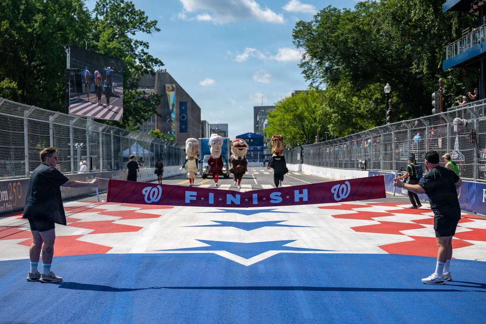 The image shows the Washington Nationals' famous 'Racing Presidents' mascots approaching a finish line banner at what appears to be a street racing or Grand Prix event. Two staff members hold a red 'FINISH' banner bearing the Washington Nationals 'W' logo across a colorfully painted track surface. The event appears to be held on a temporary street circuit lined with safety fencing and grandstands.