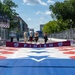 The image shows the Washington Nationals' famous 'Racing Presidents' mascots approaching a finish line banner at what appears to be a street racing or Grand Prix event. Two staff members hold a red 'FINISH' banner bearing the Washington Nationals 'W' logo across a colorfully painted track surface. The event appears to be held on a temporary street circuit lined with safety fencing and grandstands.