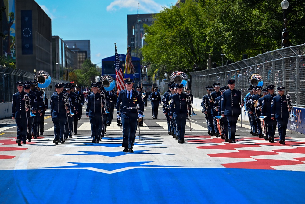 A military marching band, identifiable by dark blue Air Force dress uniforms, performs on a decorated race circuit street. The band is marching down what appears to be a Formula 1 or IndyCar Grand Prix track, indicated by the colorful track markings and safety barriers. The procession includes flag bearers carrying the American flag and a military unit flag, along with numerous brass and wind instrument players.