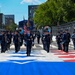 A military marching band, identifiable by dark blue Air Force dress uniforms, performs on a decorated race circuit street. The band is marching down what appears to be a Formula 1 or IndyCar Grand Prix track, indicated by the colorful track markings and safety barriers. The procession includes flag bearers carrying the American flag and a military unit flag, along with numerous brass and wind instrument players.