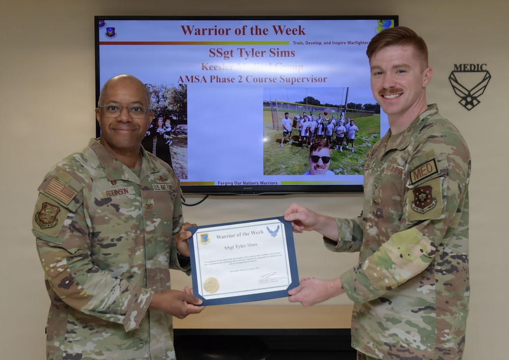 Two U.S. Air Force personnel are posing together holding a 'Warrior of the Week' certificate awarded to SSgt Tyler Sims, who serves as an AMSA Phase 2 Course Supervisor at Keesler Medical Group. The presentation is taking place in front of a display screen showing the award slide with photos of the recipient. The individual on the right appears to be SSgt Tyler Sims receiving the award from a higher-ranking officer identified by the name tape 'Robinson' on the left.
