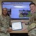 Two U.S. Air Force personnel are posing together holding a 'Warrior of the Week' certificate awarded to SSgt Tyler Sims, who serves as an AMSA Phase 2 Course Supervisor at Keesler Medical Group. The presentation is taking place in front of a display screen showing the award slide with photos of the recipient. The individual on the right appears to be SSgt Tyler Sims receiving the award from a higher-ranking officer identified by the name tape 'Robinson' on the left.