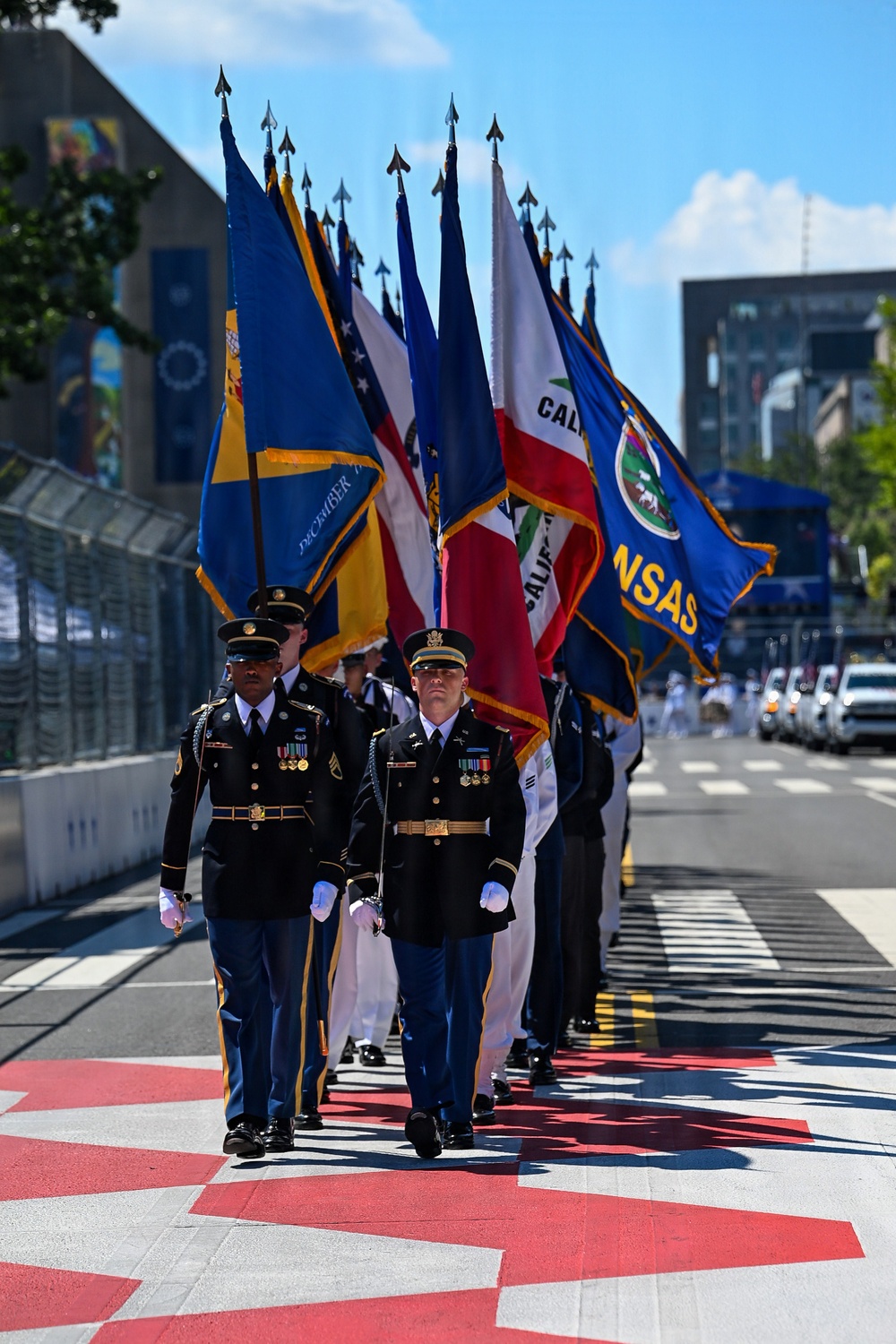 A military color guard is marching in formation along what appears to be a racing circuit or urban street course, carrying multiple state and military flags. The procession includes personnel in U.S. Army dress uniforms and Navy whites, bearing flags including California and Kansas state flags among others. The ceremony is taking place on a checkered-patterned surface typical of motorsport venues.
