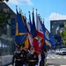 A military color guard is marching in formation along what appears to be a racing circuit or urban street course, carrying multiple state and military flags. The procession includes personnel in U.S. Army dress uniforms and Navy whites, bearing flags including California and Kansas state flags among others. The ceremony is taking place on a checkered-patterned surface typical of motorsport venues.