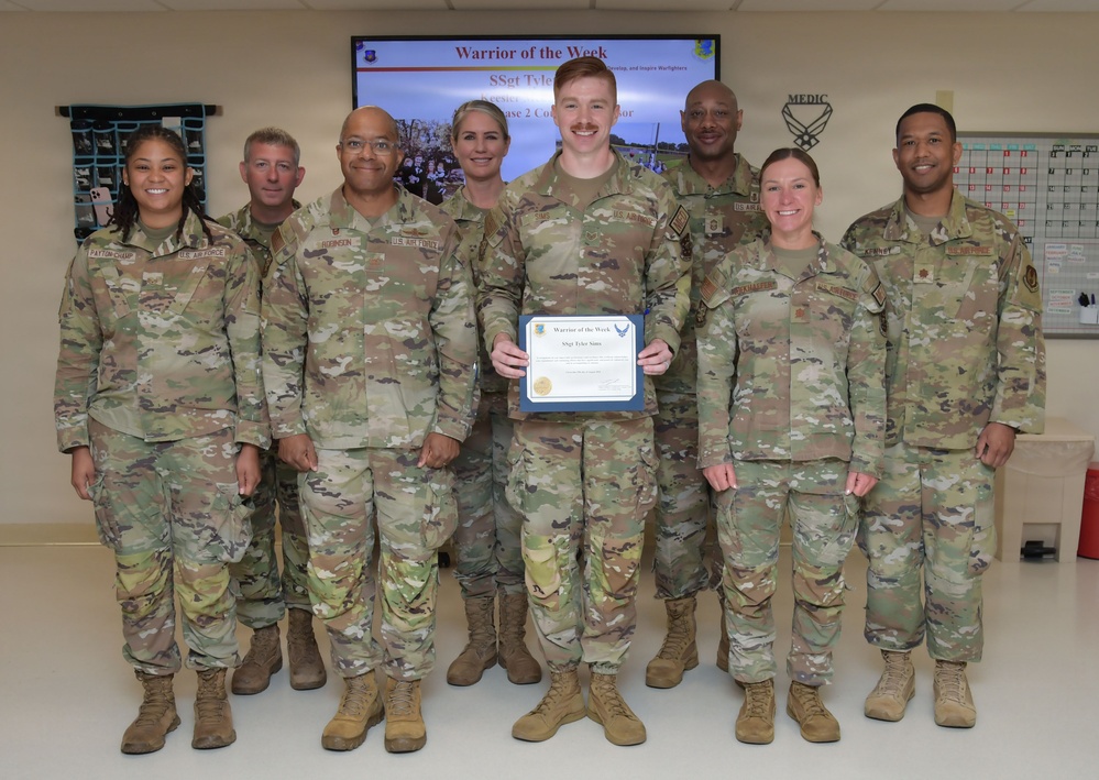 A group of eight U.S. Air Force personnel in OCP (Operational Camouflage Pattern) uniforms pose together in an indoor military facility. The central figure holds a blue-framed certificate for a 'Warrior of the Week' award, as indicated by the presentation slide visible on the screen behind them. The recipient appears to be SSgt Tyler Sims, recognized for notable performance or contribution.