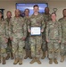 A group of eight U.S. Air Force personnel in OCP (Operational Camouflage Pattern) uniforms pose together in an indoor military facility. The central figure holds a blue-framed certificate for a 'Warrior of the Week' award, as indicated by the presentation slide visible on the screen behind them. The recipient appears to be SSgt Tyler Sims, recognized for notable performance or contribution.