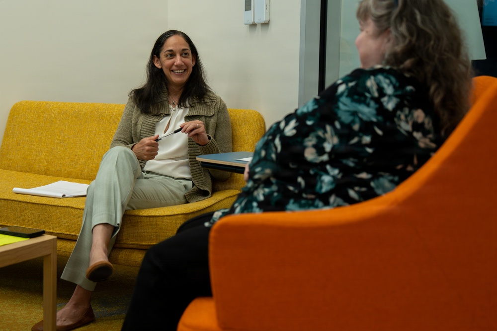 Two women are engaged in a conversation in what appears to be a modern office or lounge setting. One woman is seated on a yellow sofa, smiling and holding a pen, while the other sits in an orange chair with her back partially to the camera. The setting suggests a professional meeting, interview, or counseling session.