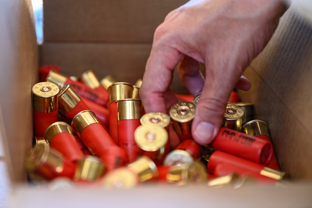 A human hand is reaching into an open cardboard box filled with numerous red shotgun shells with brass bases. The shells are loosely piled and oriented in various directions. At least one shell is partially visible with readable text, suggesting a commercial ammunition brand.