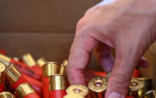 A human hand is reaching into an open cardboard box filled with numerous red shotgun shells with brass bases. The shells are loosely piled and oriented in various directions. At least one shell is partially visible with readable text, suggesting a commercial ammunition brand.