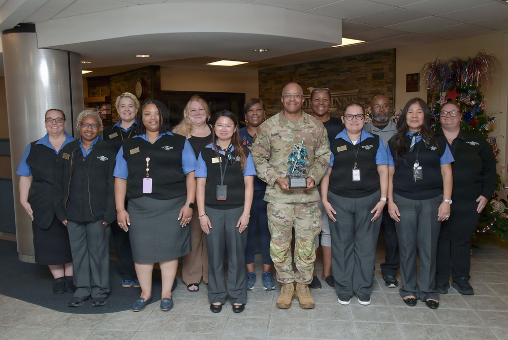 A group of approximately 13 people pose for a formal group photo in what appears to be a hotel or hospitality facility lobby. A person in U.S. Army camouflage uniform stands at the center holding a trophy or award, surrounded by staff members wearing matching black vests over blue shirts with name badges. The setting includes holiday decorations including a decorated tree visible in the background.
