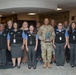 A group of approximately 13 people pose for a formal group photo in what appears to be a hotel or hospitality facility lobby. A person in U.S. Army camouflage uniform stands at the center holding a trophy or award, surrounded by staff members wearing matching black vests over blue shirts with name badges. The setting includes holiday decorations including a decorated tree visible in the background.