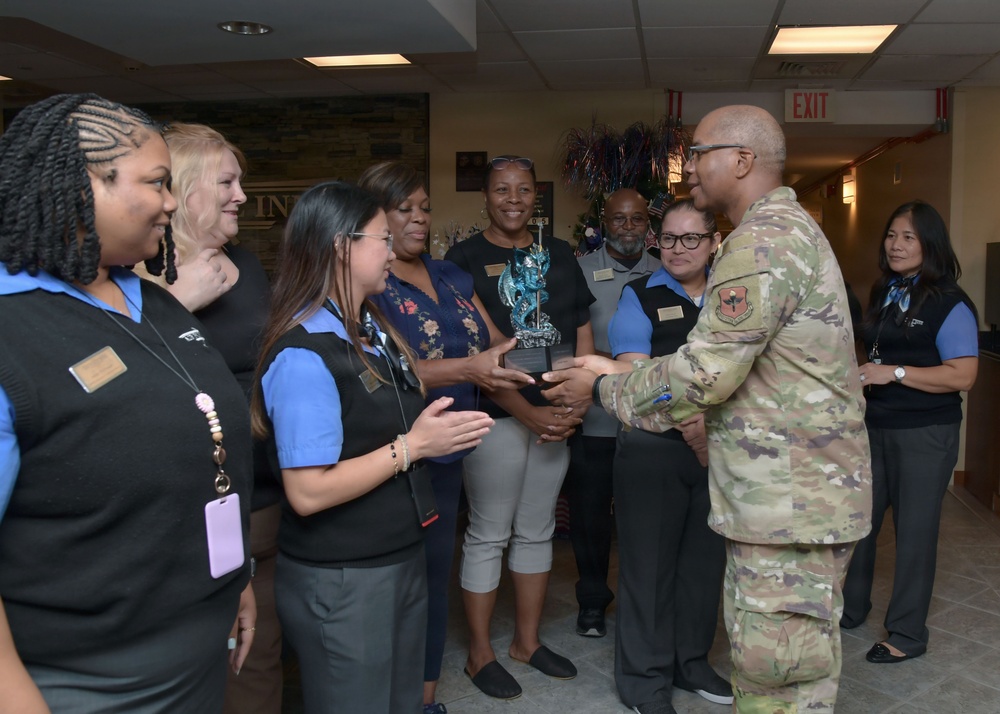 A military officer in Army Combat Uniform (ACU/OCP camouflage pattern) is presenting a decorative trophy or award to a group of civilian staff members wearing blue and black uniforms with name badges. The ceremony appears to be taking place in an indoor facility, likely a military installation service building. The group of approximately eight civilian employees is gathered to receive the recognition.