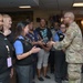 A military officer in Army Combat Uniform (ACU/OCP camouflage pattern) is presenting a decorative trophy or award to a group of civilian staff members wearing blue and black uniforms with name badges. The ceremony appears to be taking place in an indoor facility, likely a military installation service building. The group of approximately eight civilian employees is gathered to receive the recognition.