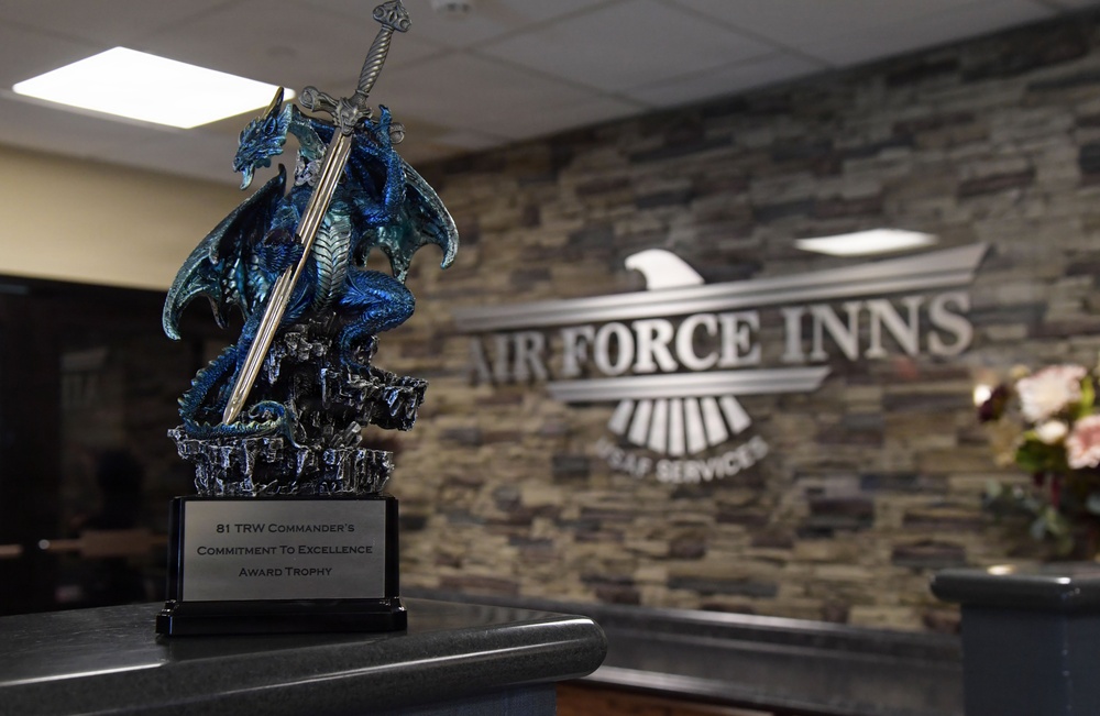 A decorative dragon trophy holding a sword sits on a counter at an Air Force Inns lodging facility. The trophy's plaque identifies it as the '81 TRW Commander's Commitment To Excellence Award Trophy.' The background features a stone-textured wall with a metallic 'Air Force Inns' sign and an eagle emblem.