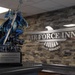 A decorative dragon trophy holding a sword sits on a counter at an Air Force Inns lodging facility. The trophy's plaque identifies it as the '81 TRW Commander's Commitment To Excellence Award Trophy.' The background features a stone-textured wall with a metallic 'Air Force Inns' sign and an eagle emblem.