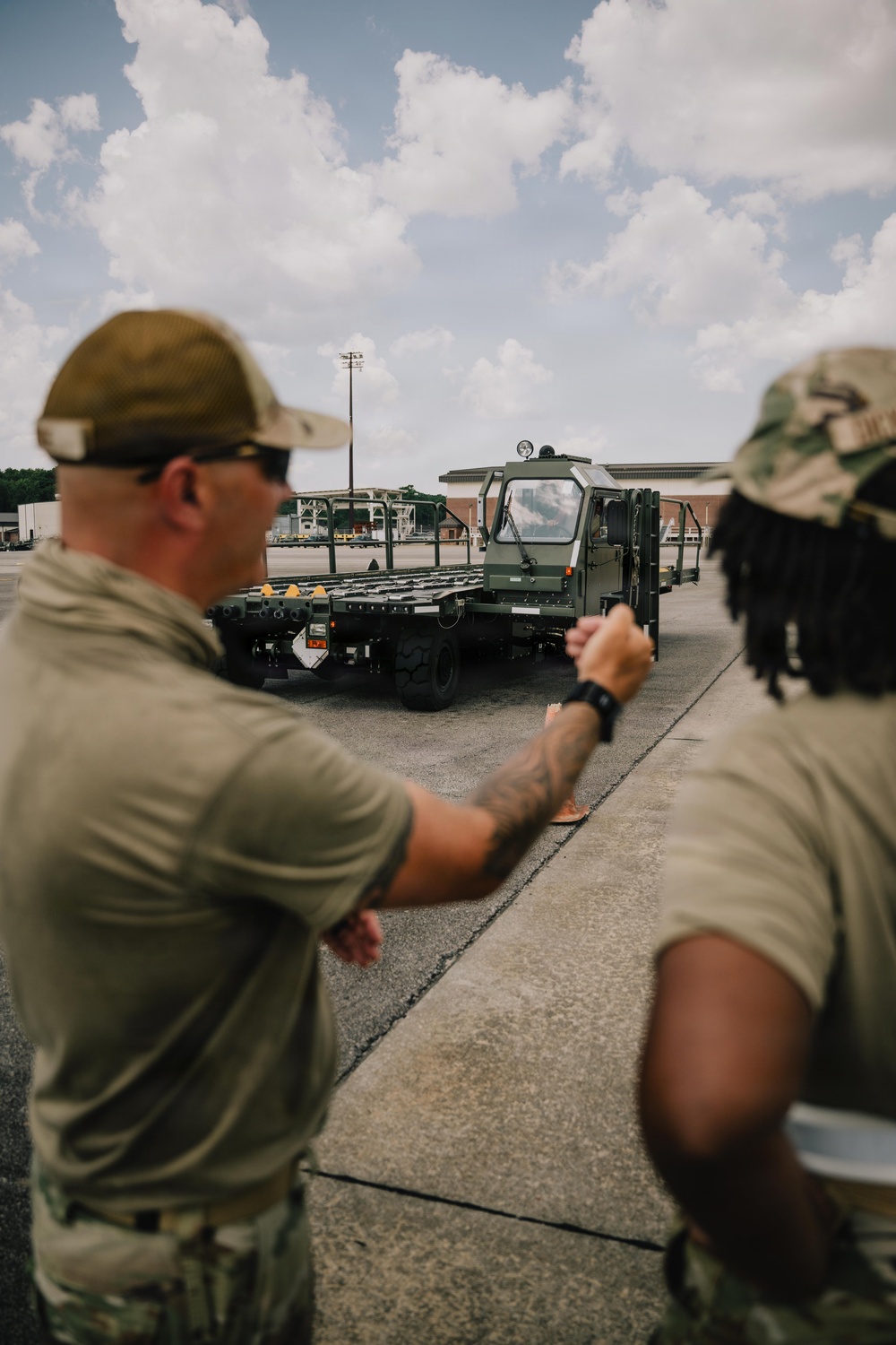 Two military personnel in olive drab/tan uniforms are observing a military flatbed vehicle on a paved surface, likely an airfield or logistics yard. One individual appears to be gesturing or directing toward the vehicle. The setting suggests a training or operational logistics activity.