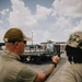Two military personnel in olive drab/tan uniforms are observing a military flatbed vehicle on a paved surface, likely an airfield or logistics yard. One individual appears to be gesturing or directing toward the vehicle. The setting suggests a training or operational logistics activity.
