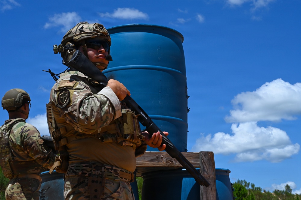 Two military personnel in full combat gear are positioned near large blue barrels in an outdoor training or operational environment. The primary soldier in the foreground is equipped with a tactical helmet, body armor, and is holding a shotgun in a ready position. The setting appears to be a military training exercise based on the controlled environment and equipment visible.
