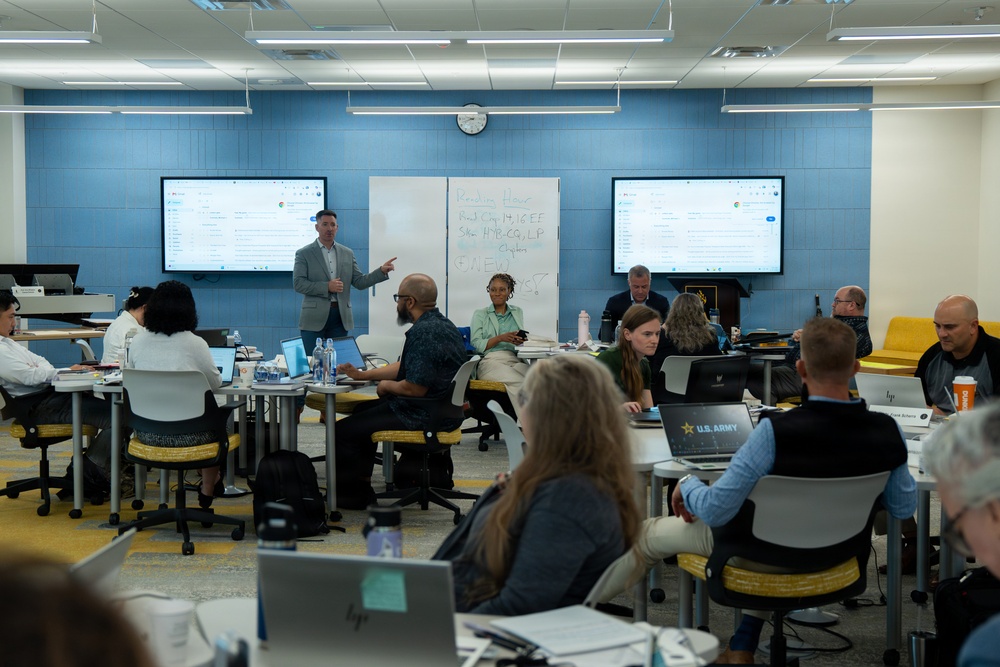 A professional training or workshop session is taking place in a modern classroom or conference room with blue accent walls. A male presenter in a gray blazer stands at the front, gesturing toward a whiteboard while participants seated at multiple tables work on laptops. At least one laptop displays a 'U.S. Army' sticker, suggesting a military-affiliated or government training context.
