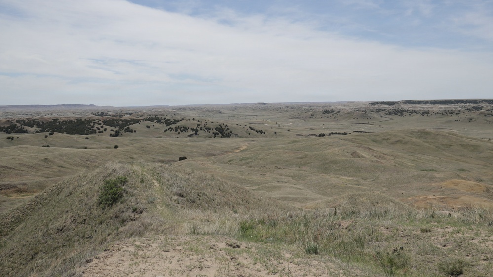 A wide panoramic view of a semi-arid grassland landscape featuring rolling hills and scattered vegetation. The terrain appears to be characteristic of the Great Plains or badlands region, with sparse grass cover, scattered shrubs and trees, and eroded hillsides. The sky is partly cloudy with a flat horizon visible in the far distance.