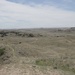 A wide panoramic view of a semi-arid grassland landscape featuring rolling hills and scattered vegetation. The terrain appears to be characteristic of the Great Plains or badlands region, with sparse grass cover, scattered shrubs and trees, and eroded hillsides. The sky is partly cloudy with a flat horizon visible in the far distance.