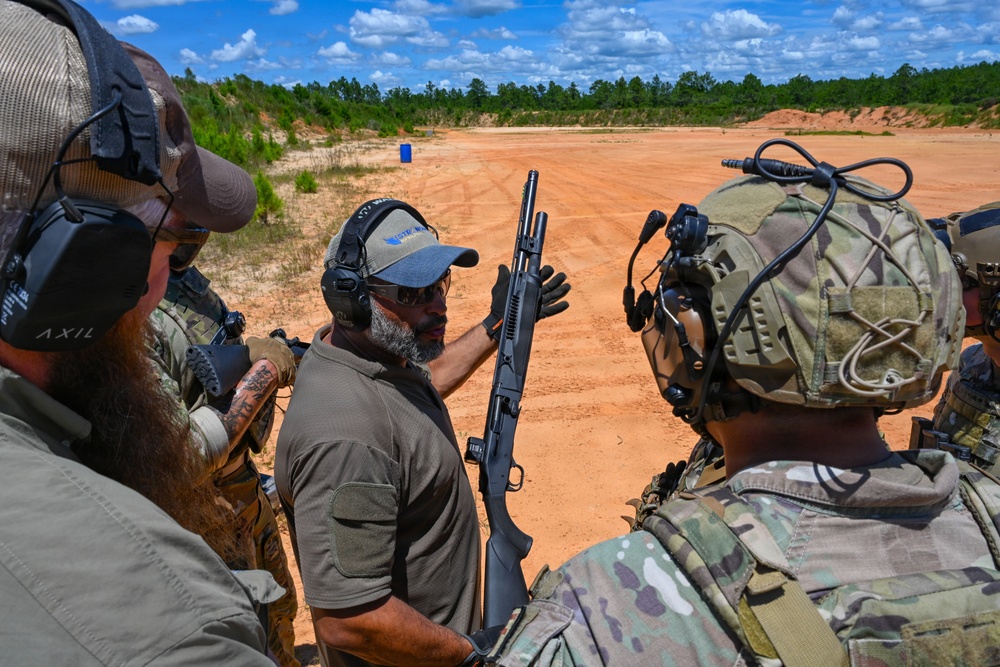 An instructor in civilian attire is demonstrating a pump-action shotgun to a group of military personnel in full tactical gear at an outdoor shooting range. The setting appears to be a formal firearms training exercise conducted on a sandy range with target markers visible in the background. The participants are all wearing hearing protection, indicating active range safety protocols.