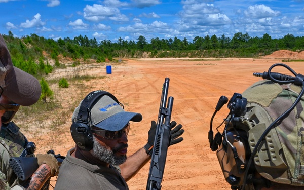 An instructor in civilian attire is demonstrating a pump-action shotgun to a group of military personnel in full tactical gear at an outdoor shooting range. The setting appears to be a formal firearms training exercise conducted on a sandy range with target markers visible in the background. The participants are all wearing hearing protection, indicating active range safety protocols.