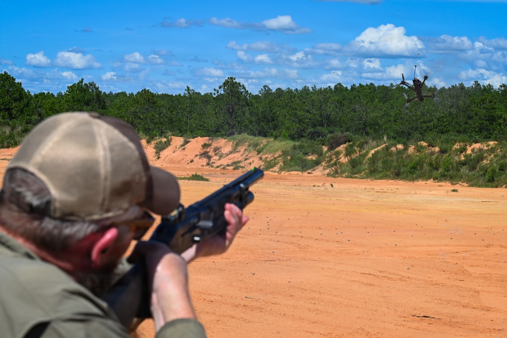 A person at an outdoor shooting range is aiming a shotgun at a drone in mid-air. The drone appears to have been struck and is tumbling/falling in the background. The setting is a sandy open range area surrounded by pine trees under a partly cloudy sky.