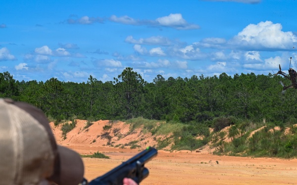 A person at an outdoor shooting range is aiming a shotgun at a drone in mid-air. The drone appears to have been struck and is tumbling/falling in the background. The setting is a sandy open range area surrounded by pine trees under a partly cloudy sky.