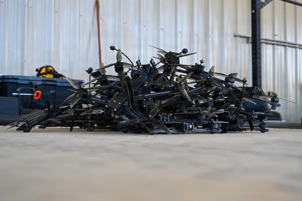 A large pile of disassembled or damaged quadcopter/multirotor drones is stacked on a concrete floor inside what appears to be a warehouse or hangar. The drones are predominantly black and appear to be small consumer or commercial-grade UAVs with visible propeller blades, frames, and landing gear. The pile suggests either a collection of seized, crashed, or decommissioned drones.