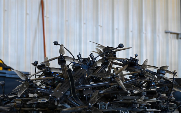 A large pile of disassembled or damaged quadcopter/multirotor drones is stacked on a concrete floor inside what appears to be a warehouse or hangar. The drones are predominantly black and appear to be small consumer or commercial-grade UAVs with visible propeller blades, frames, and landing gear. The pile suggests either a collection of seized, crashed, or decommissioned drones.