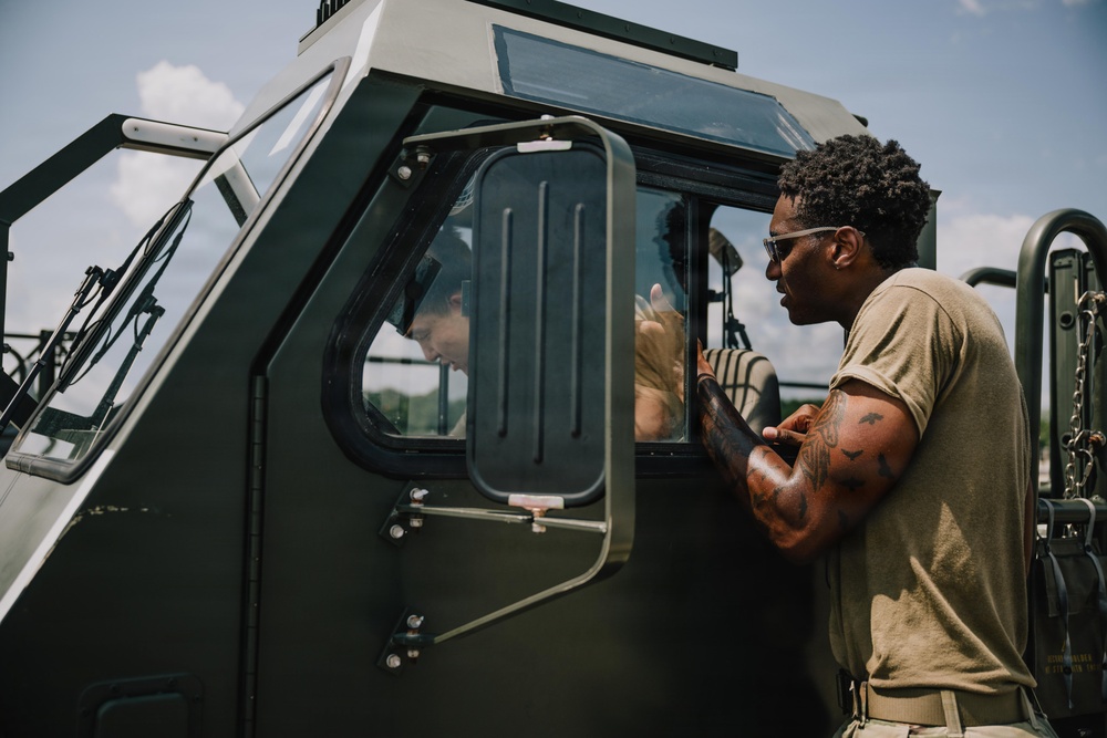 Two individuals in olive/tan military-style clothing are interacting beside a dark green military vehicle. One person stands outside the vehicle leaning toward the open window, while another is seated inside the cab. The setting appears to be an outdoor military or training environment under a partly cloudy sky.