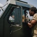 Two individuals in olive/tan military-style clothing are interacting beside a dark green military vehicle. One person stands outside the vehicle leaning toward the open window, while another is seated inside the cab. The setting appears to be an outdoor military or training environment under a partly cloudy sky.