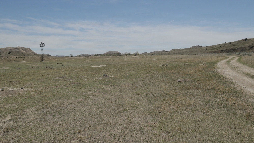A wide open semi-arid grassland landscape featuring sparse, dry vegetation and eroded badlands-style hills in the background. A traditional farm windmill stands on the left side of the frame, and a dirt two-track road curves along the right side. The scene suggests a rural, undeveloped rangeland environment, likely in the Great Plains region of the United States.