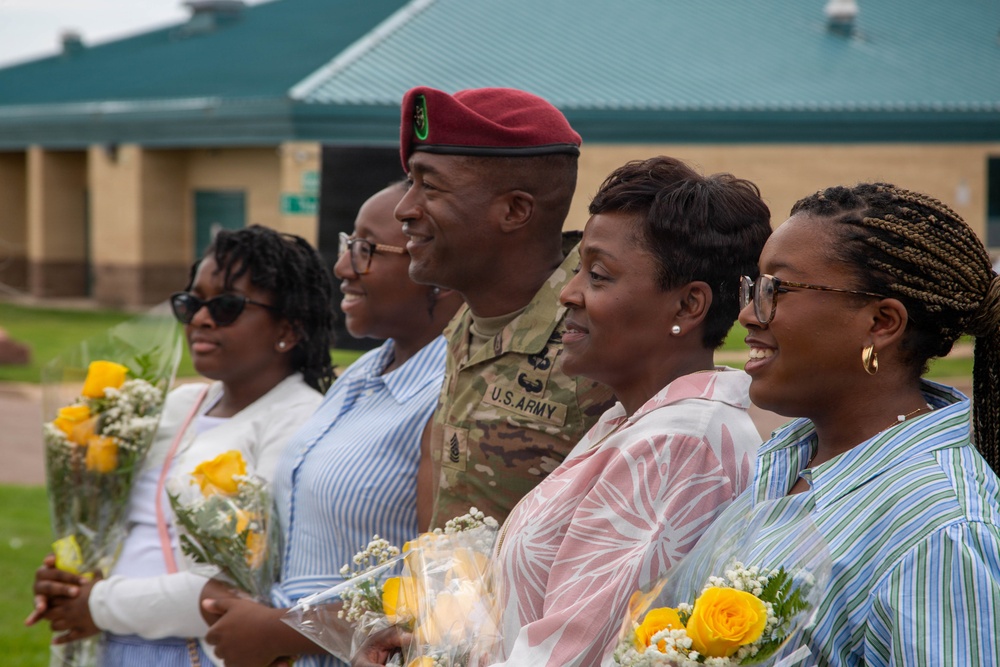 A U.S. Army soldier in camouflage uniform and maroon beret poses with four civilian women holding yellow rose bouquets. The group appears to be at a military ceremony or celebration, likely a homecoming, promotion, or retirement event. All individuals are smiling and appear in good spirits in an outdoor setting near a military building.