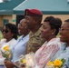 A U.S. Army soldier in camouflage uniform and maroon beret poses with four civilian women holding yellow rose bouquets. The group appears to be at a military ceremony or celebration, likely a homecoming, promotion, or retirement event. All individuals are smiling and appear in good spirits in an outdoor setting near a military building.