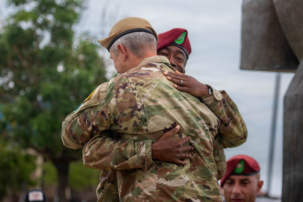 Two military personnel in camouflage uniforms are embracing in what appears to be a formal or ceremonial setting. One individual wears a tan/khaki beret and the other wears a maroon/red beret with a green flash insignia. A third military person wearing a matching red beret is partially visible in the background foreground.