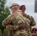 Two military personnel in camouflage uniforms are embracing in what appears to be a formal or ceremonial setting. One individual wears a tan/khaki beret and the other wears a maroon/red beret with a green flash insignia. A third military person wearing a matching red beret is partially visible in the background foreground.
